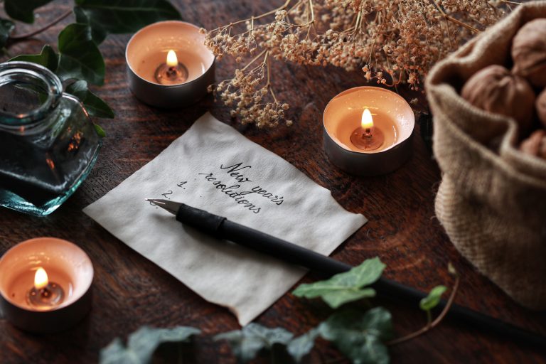 White peace of paper with "New Year's Resolutions" written on it. Yule winter solstice (Christmas) theme with nature themed background of dried flowers ivy branch, walnuts, ink bottle, pen and candles